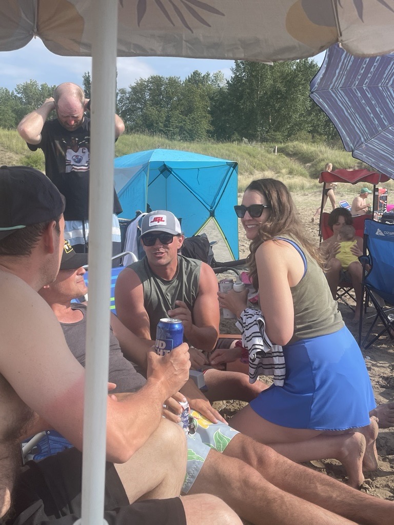 Group of people relaxing on a sandy beach under a large patterned umbrella — a woman in a green tank top holds a baby, a man in a white hat holds a blue can, a blue beach tent visible behind them