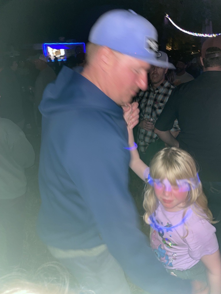 A man in a blue hoodie holding hands with a young girl wearing a light purple shirt and glowing blue neon accessories, surrounded by a festive dimly lit crowd