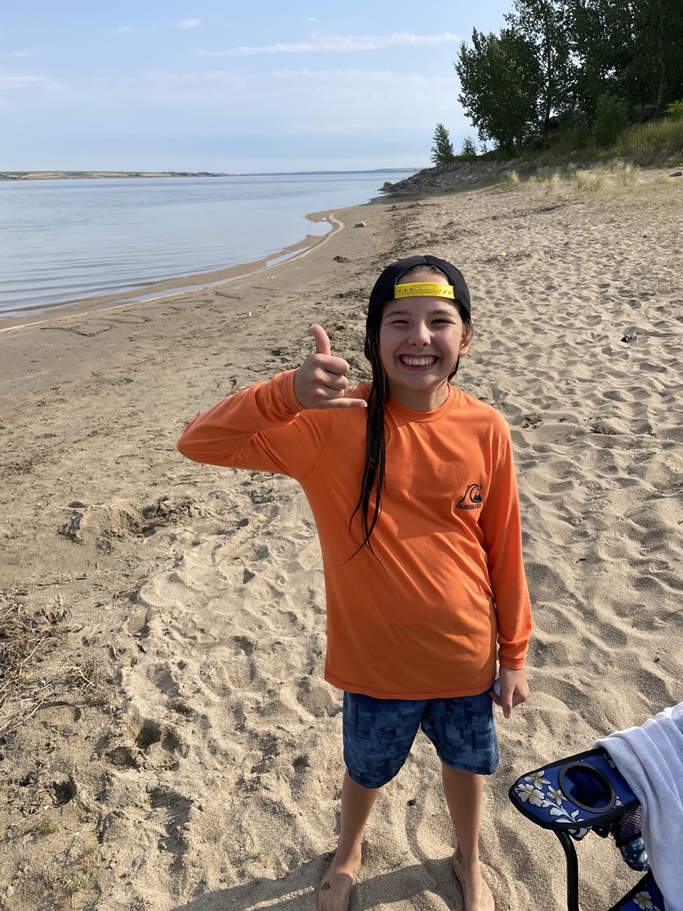 A boy with long dark hair in an orange long-sleeve shirt and black cap with yellow brim, standing on the sandy beach making a shaka hand gesture with calm blue water behind him
