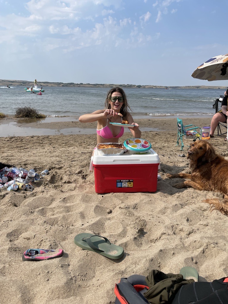 Woman in a pink bikini top and sunglasses on the sandy beach, holding food over a red Igloo cooler, a golden retriever lying relaxed beside it — prime beach day energy