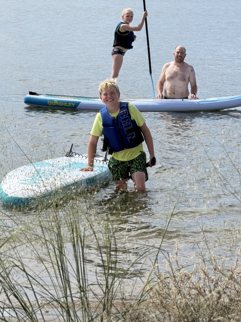 Two boys and a man in shallow lake water — a boy in a yellow life vest stands by a light blue paddleboard, another boy stands on a blue paddleboard further back, a man steadying him