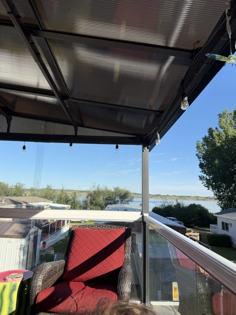 View from the covered cabin balcony — a dark red wicker chair with red cushion in the foreground, calm lake and distant houses visible beyond the glass railing under a clear blue sky
