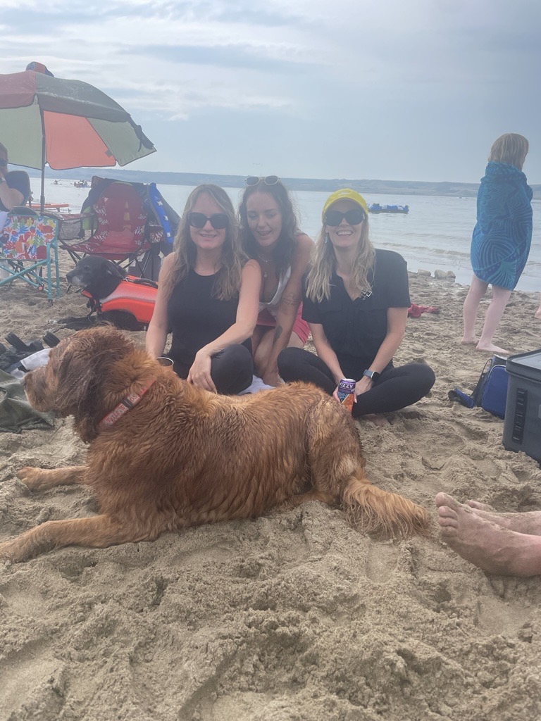 Three women and a golden retriever sitting on a sandy beach — two wear black tops and sunglasses, the third has a yellow headband, the dog lies relaxed in front of them